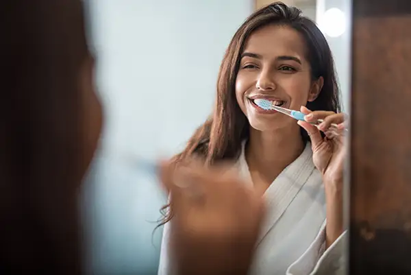 Young woman in a white robe smiling and brushing her teeth while looking into a bathroom mirror, practicing good dental hygiene.