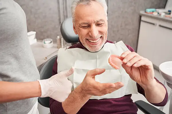 A smiling senior patient inspects his new dentures while a dental assistant helps him with it.